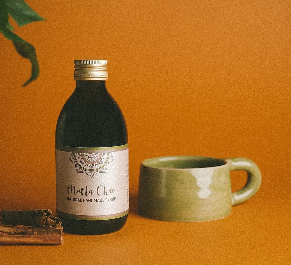 Chai-Sirup-Flasche mit Zimtstangen, gruener Tasse und Pflanzenblatt auf leuchtend orangenem Hintergrund
